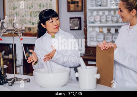 Japanische Apothekerin macht Handgesten im Stehen und spricht mit einer kaukasischen Kollegin, die die Inhaltsstofftabelle in der Vintage-Apotheke hält Stockfoto