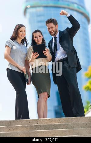 Lächelnde asiatische Geschäftsfrau, die ein Tablet benutzt, mit kaukasischen Kollegen, die an sonnigen Tagen auf einer Außentreppe stehen Stockfoto