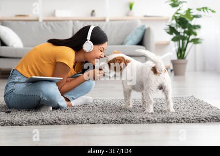 Asiatische Frau mit Kopfhörern kuschelt Hund auf dem Boden mit Teppich im Wohnzimmer zu Hause Stockfoto