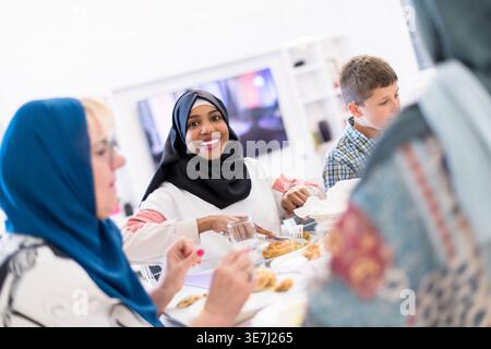 Lächelnde schwarze Frau im Hijab mit kaukasischen Freunden, die zusammen essen und zu Hause am Tisch sitzen Stockfoto