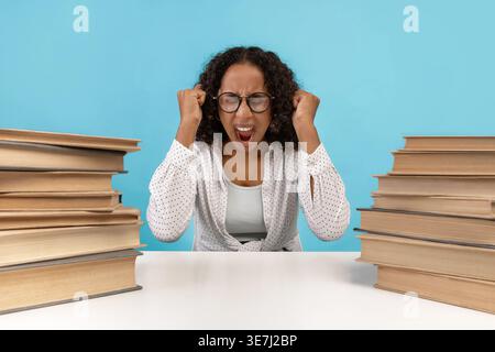 Gestresste schwarze Teenagerin mit Brille und schreiender Brille, während sie im Studio mit Bücherstapeln vor blauem Hintergrund am Tisch sitzt Stockfoto