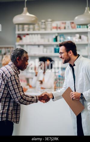 Senior Black man schüttelt die Hand mit einem männlichen kaukasier, der ein Klemmbrett hält, der in der Apotheke gegen den Tresen steht Stockfoto