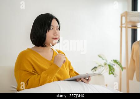 Chinesische Frau in Übergröße, die Stifte und Tablets hält und wegblickt, während sie zu Hause auf dem Sofa sitzt Stockfoto
