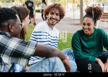 Fröhliche, abwechslungsreiche Gruppe von Freunden, die tagsüber zusammen auf Gras im Park sitzen und lachen Stockfoto