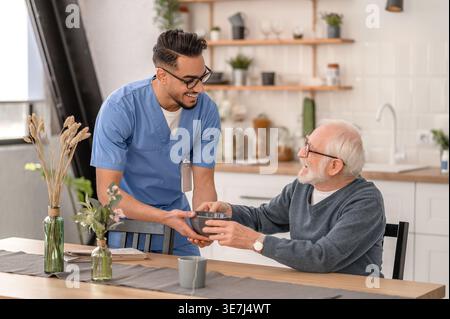Lächelnder arabischer männlicher Pfleger mit Brille, der Essen in der Nähe eines älteren Mannes gibt, während er zu Hause am Tisch sitzt Stockfoto