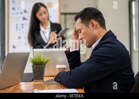 Gestresster asiatischer Geschäftsmann, der die Stirn mit geschlossenen Augen berührt und Probleme mit seiner Arbeit hat, während er am Tisch im Büro sitzt Stockfoto