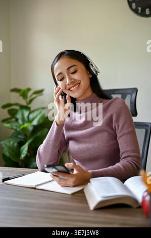 Lächelnde asiatische Frau im Kopfhörer, die Musik mit dem Smartphone hört, während sie zu Hause mit Büchern am Tisch sitzt Stockfoto