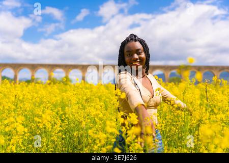Lächelnde Schwarze Frau mit Wendungen, die mit ausgestreckten Armen im Feld der gelben Blumen an sonnigem Tag steht Stockfoto
