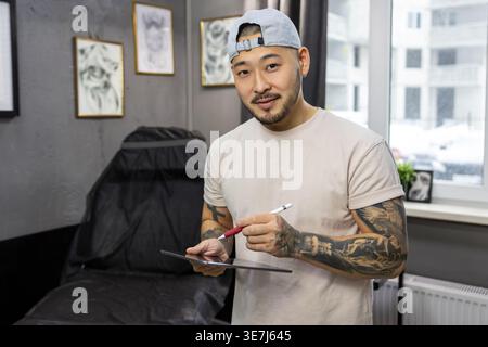 Ein lächelnder asiatischer Tätowierer, der auf Tablet neue Kunstideen ausarbeitet, während er in seinem Tätowierstudio steht Stockfoto