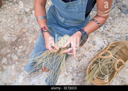 Hispanische Frau mit Denimschürze, die auf einem kleinen Stuhl sitzt und tagsüber einen Korb mit Espartofasern vor ihrem Geschäft webt Stockfoto
