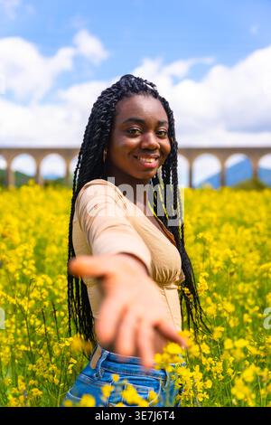 Lächelnde schwarze Frau mit Wendungen, die mit offenen Händen im Feld der gelben Blumen an sonnigem Tag steht Stockfoto