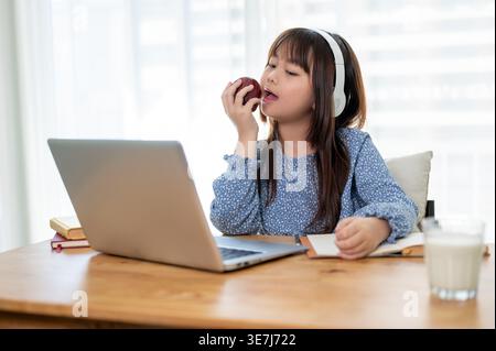 Asiatisches Mädchen, das Kopfhörer trägt und Apfel isst, während er zu Hause auf den Laptop blickt Stockfoto