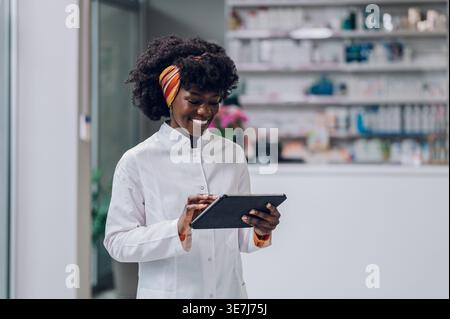 Schwarze Apothekerin mit Afro-Haltung und Verwendung der Tablette, die in der Apotheke gegen den Zähler steht Stockfoto