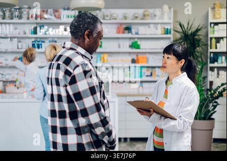 Japanische Apothekerin schreibt auf die Zwischenablage und spricht mit einem älteren schwarzen männlichen Kunden, der in der Apotheke steht Stockfoto