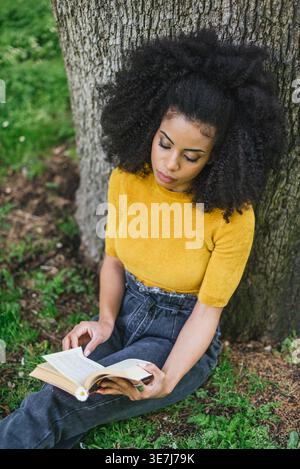 Ernste hispanische Frau mit lockigem Haar, die tagsüber auf dem Boden sitzt und ein Buch liest, das sich im Park gegen den Baumstamm lehnt Stockfoto