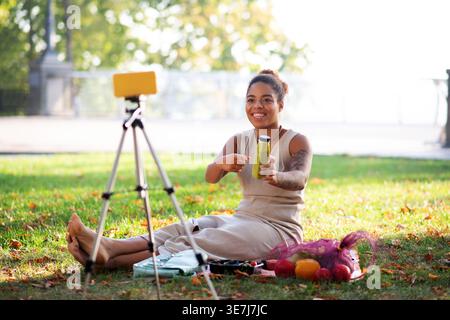 Lächelnde Mixed Race Frau, die auf eine Flasche gesunder Smoothie-Aufnahme zeigt, während sie am sonnigen Tag auf der Matte im Park sitzt Stockfoto