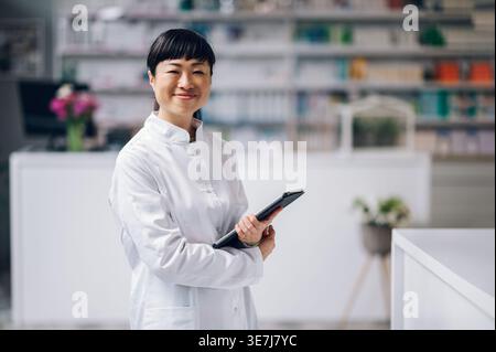 Fröhliche japanische Apothekerin mit Tablette im Arm, die in der Apotheke gegen den Zähler steht Stockfoto