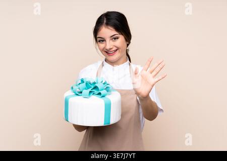 Hispanische Konditorin zeigt Handgeste, die Kuchen im Studio vor beigefarbenem Hintergrund hält Stockfoto
