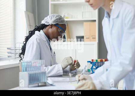 Ernste schwarze Medizintechnikerin, die Laborkittel mit roter Substanz trägt und in der Nähe einer kaukasischen Kollegin in der Medizin ... sitzt Stockfoto