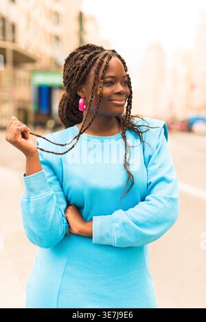 Lächelnde schwarze Frau mit Wendungen, die blauen Pullover trägt und wegblickt, während sie auf der Stadtstraße steht Stockfoto