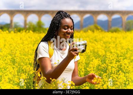 Lächelnde schwarze Frau mit Wendungen, die gelbe Blumen fotografiert, die am sonnigen Tag auf dem Feld stehen Stockfoto