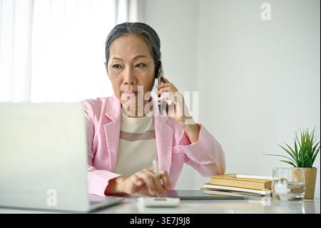 Lächelnde ältere asiatische Frau, die telefoniert und den Laptop anschaut, während sie im Home Office am Tisch sitzt Stockfoto