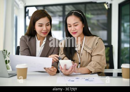 Lächelnde asiatische Buchhalterinnen arbeiten und analysieren Finanzdatenberichte, während sie zusammen am Tisch im Büro sitzen Stockfoto