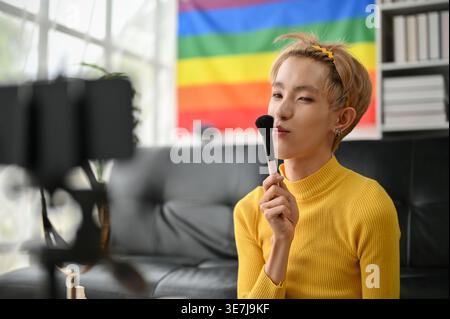 Lächelnder asiatischer schwuler Mann Aufnahme Make Up Tutorial zeigt Make-up Pinsel auf der Kamera, während Sie auf dem Boden in der Nähe des Sofas zu Hause sitzen Stockfoto