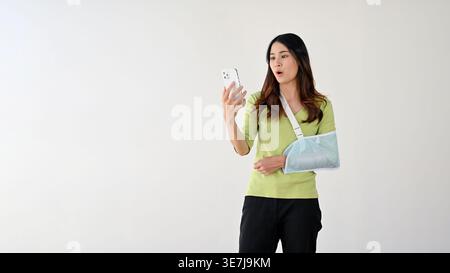 Staunte asiatische Frau, die einen Armschlinge mit einem Smartphone trägt, das vor grauem Hintergrund steht Stockfoto