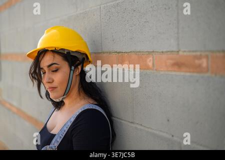 Hispanische Arbeiterin Denim-Overall und gelbem Hardhat im Stehen mit geschlossenen Augen an der grauen Wand auf der Baustelle... Stockfoto