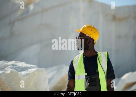 Schwarzer männlicher Ingenieur mit Schutzhelm und Sonnenbrille, der wegblickt, während er auf der Baustelle steht Stockfoto