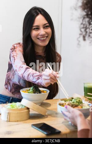 Gemischte Frau, die Essen mit Essstäbchen mit einer hispanischen Freundin teilt, während sie am Holztisch im Restaurant sitzt Stockfoto