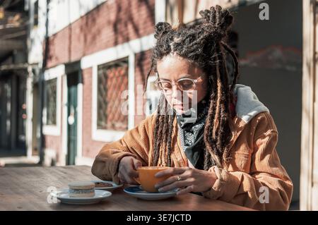 Hispanische Frau mit Dreadlocks, die eine Brille trägt, die eine Kaffeetasse hält und tagsüber am Tisch im Café im Freien sitzt Stockfoto