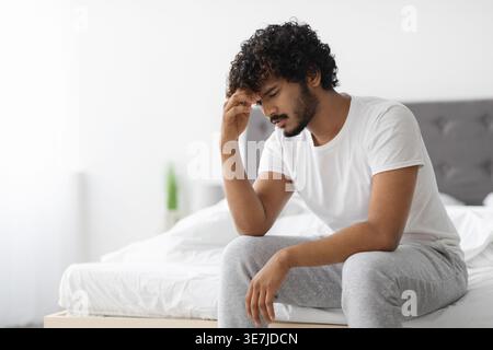 Ernsthafter bärtiger Indianer mit lockigem Haar, der die Stirn berührt, sitzt auf dem Bett zu Hause Stockfoto