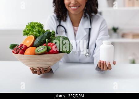 Zugeschnittenes Bild eines schwarzen weiblichen Ernährungsberaters, der eine Schüssel mit Obst und Gemüse und eine Flasche Pillen hält, während er in der Klinik am Tisch sitzt Stockfoto