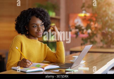 Schwarze Frau mit lockigem Haar, die Notizen schreibt, sitzt am Tisch mit Smartphone und Laptop im Café Stockfoto
