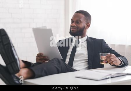 Schwarzer Geschäftsmann mit Dokumenten und einem Glas Whiskey, der im Büro mit den Beinen nach oben auf dem Tisch sitzt Stockfoto