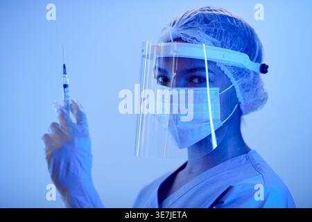 Gemischte Krankenschwester mit Schutzmaske und OP-Kappe, die die medizinische Injektion seitlich im blau beleuchteten Labor hält Stockfoto