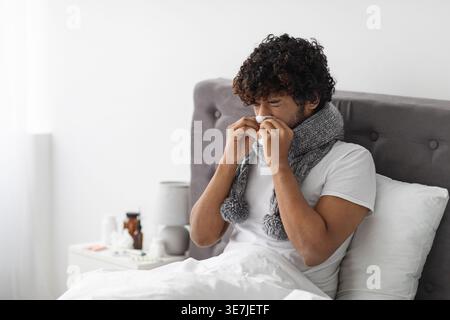 Ein kranker asiatischer Mann mit lockigen Haaren, der einen Schal um den Hals trägt und sich mit einem Taschentuch die Nase abwischt, während er zu Hause im Bett sitzt Stockfoto