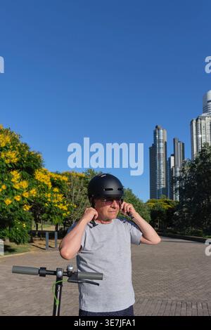 Senior hispanischer Mann mit Sonnenbrille, der Helm steht mit Elektroroller im Stadtpark an sonnigen Tagen Stockfoto