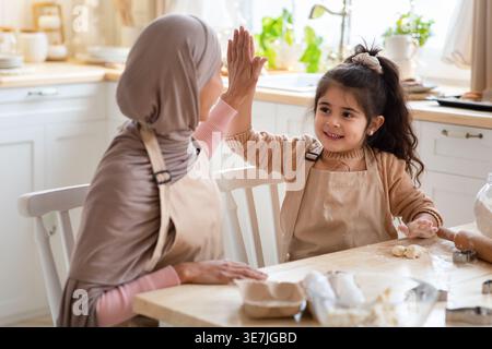 Mutter aus dem Nahen Osten im Hijab und Tochter mit Schürzen, die sich gegenseitig hohe fünf geben, während sie am Tisch mit Zutaten in der Küche zu Hause sitzt Stockfoto