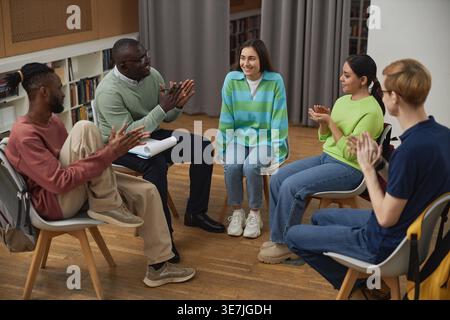 Schwarzer männlicher Psychologe und eine Gruppe von Kollegen klatschen die Hände für eine kaukasische Patientin, während sie während der Beratung auf dem Stuhl saßen... Stockfoto