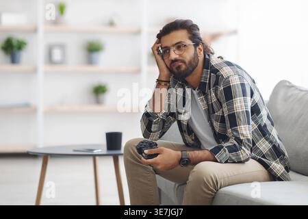 Gelangweilter arabischer Mann, der eine Brille trägt und den Gamecontroller hält und wegblickt, während er auf dem Sofa im Wohnzimmer sitzt Stockfoto