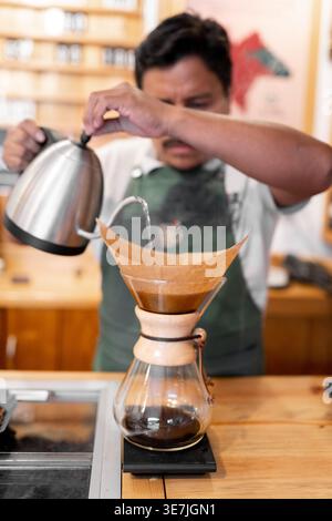 Männlicher Barista, der heißes Wasser für gefilterten Kaffee gießt, während er am Tisch im Café steht Stockfoto