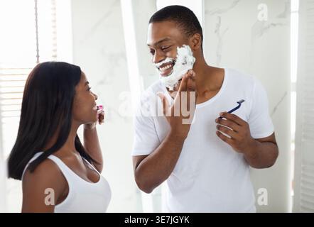 Schwarzer Freund, der Rasiercreme auf das Gesicht aufträgt und die Freundin mit einer Zahnbürste ansieht, während er zu Hause im Badezimmer steht Stockfoto