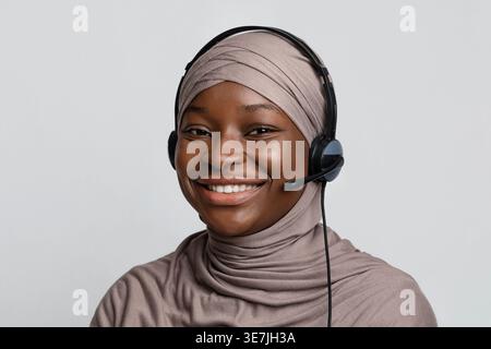 Schwarze muslimische Callcenter-Agentin im Hijab mit Headset, die im Studio vor grauem Hintergrund steht Stockfoto