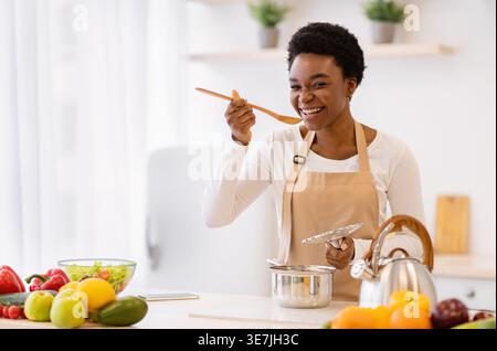 Schwarze Frau mit kurzen Haaren, die Schürze trägt, Kochen und Verkochen mit Holzlöffel, während sie zu Hause in der Küche steht Stockfoto