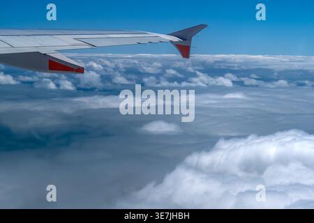 Blick von einem Passagierflugzeugfenster aus, das den Flugzeugflügel mit roten Akzenten zeigt, der über einer dramatischen Schicht weißer Cumulus-Wolken unter einer lebendigen Decke aufragt Stockfoto