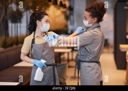 Gemischte und asiatische Kellnerinnen mit Gesichtsmasken und Schürzen, die sich gegenseitig am Ellenbogen stoßen, während sie während der Pandemie im Café stehen Stockfoto