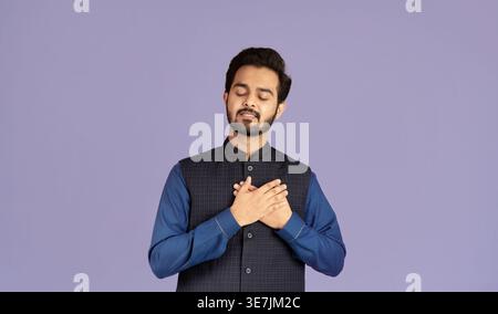 Indischer Mann mit geschlossenen Augen, der Hände in der Nähe des Herzens hält und vor violettem Hintergrund im Studio steht Stockfoto